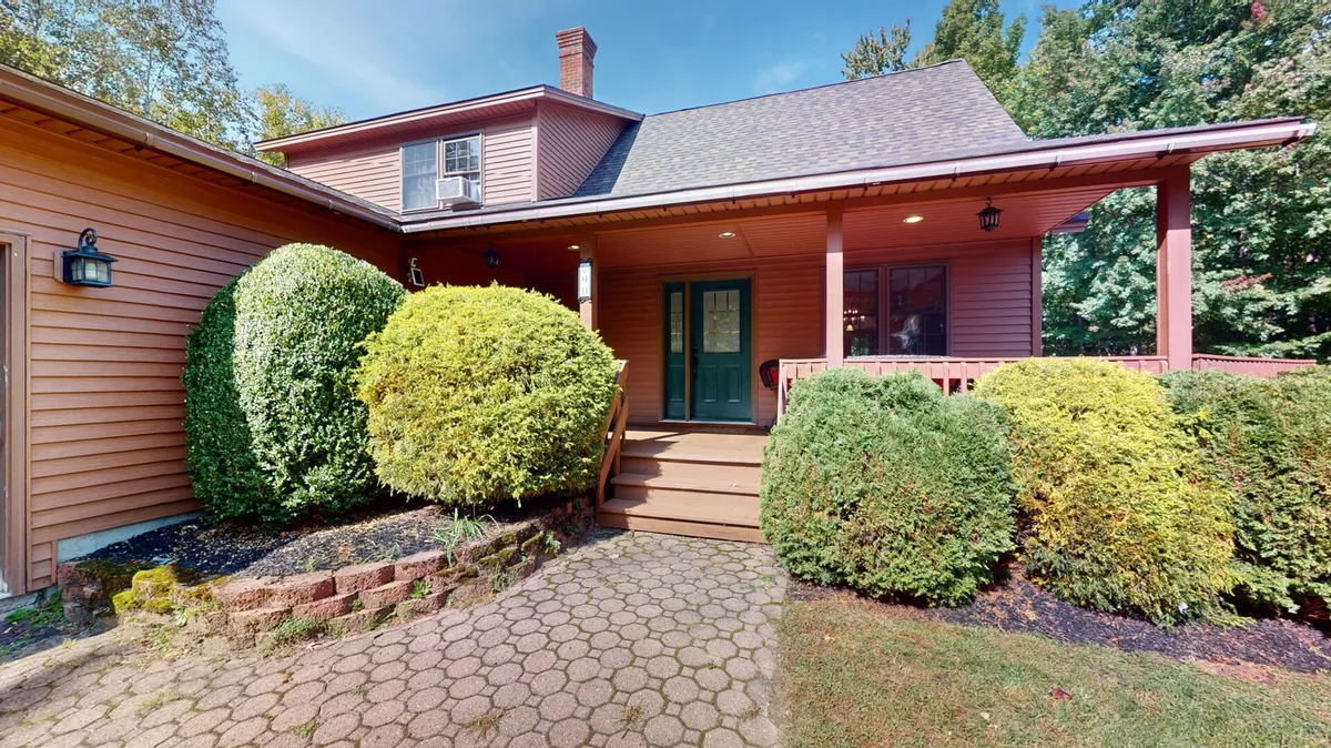 This image captures the home's front porch framed by lush green shrubs and wooden steps leading to a warm doorway. The inviting porch area subtly communicates the presence of exterior security cameras, a welcoming entry point, and the seamless indoor-outdoor flow important for year-round living.