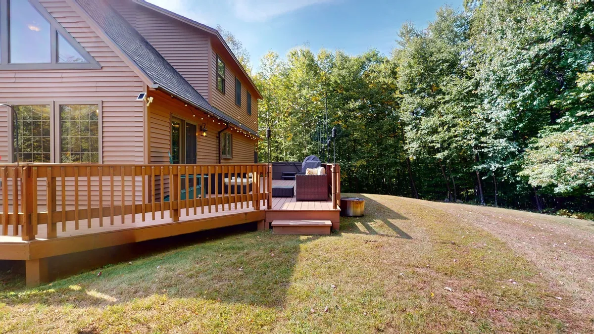 The image shows a spacious back deck with comfortable seating surrounded by lush Vermont trees. The area is designed for relaxation and gatherings, offering views of the forest and an inviting atmosphere for outdoor activities. The deck serves as a seamless extension of the home's interior, with natural wood elements and ample space for enjoying the outdoors.