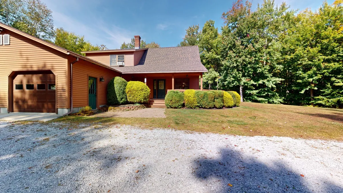 This image captures the front porch of the home, surrounded by lush greenery, a gravel driveway, and a visible garage. The scene exudes a welcoming outdoor atmosphere, with natural foliage and an inviting space for relaxation or gatherings, suggesting easy access to Vermont's scenic outdoors.