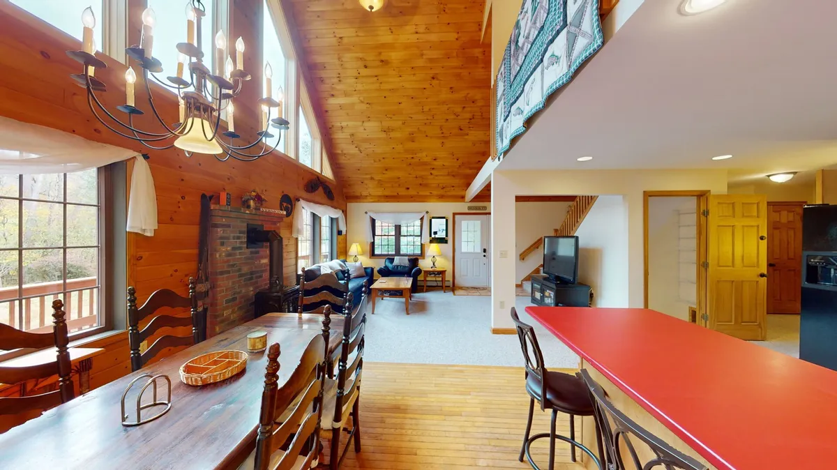 A large dining table sits under a chandelier, adjacent to a fireplace and the red kitchen counter. Wooden beams create a warm, welcoming atmosphere that fosters memorable family meals and celebrations.