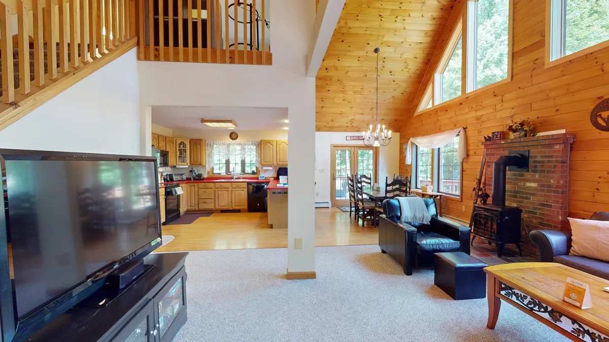 A warm, inviting common room featuring large windows, exposed wooden beams, a central fireplace, and a dining area. The space is ideal for family gatherings, relaxing after winter sports, or sharing meals year-round.