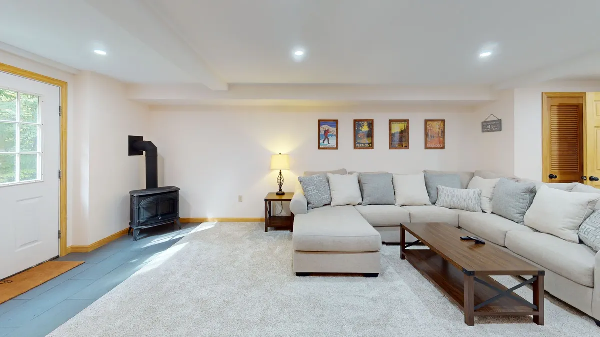 A spacious family room featuring a large sectional sofa, coffee table, floor lamp, and wall artwork. The room is brightened by natural light from a nearby door leading outside, creating a welcoming environment for gatherings, movie nights, or games.