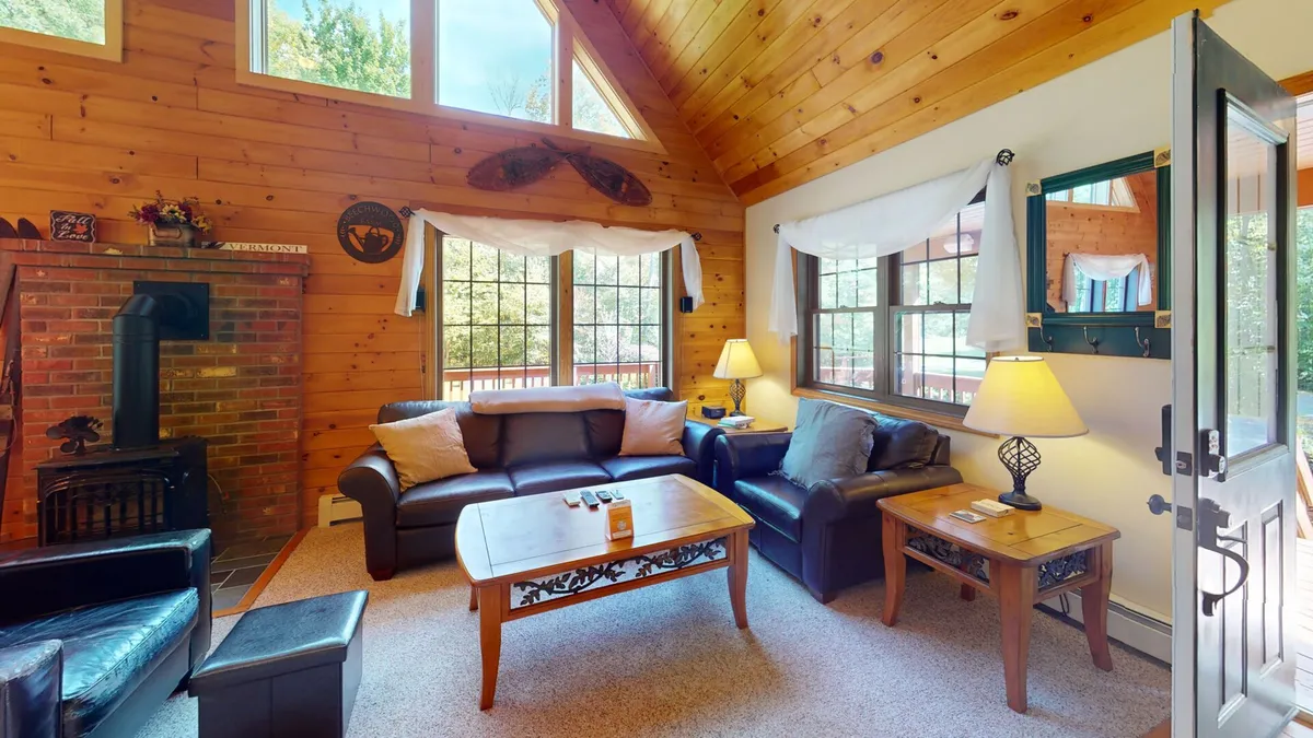 This image captures a warm living room space featuring a striking brick fireplace, expansive floor-to-ceiling windows, a wooden vaulted ceiling, plush sofas, and a coffee table. Natural light pours in, and the view of the outdoors blends seamlessly with the interior.