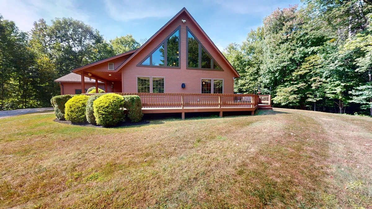 The image showcases the exterior of a spacious single-family home nestled among green woods, featuring a striking triangular window design, a large front deck perfect for gatherings, and beautifully landscaped bushes. The setting evokes a peaceful, nature-filled retreat with ample space to relax outdoors.