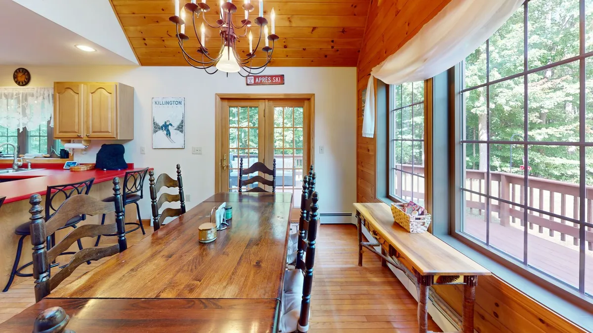 Showcasing a spacious dining area with a long wooden table, elegant chandelier, and large windows. The wooden bench and chairs welcome groups, and the natural light enhances the warm, communal atmosphere.